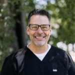 Smiling man in black scrubs with glasses outdoors, background of trees and bokeh light.