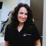 Smiling woman in black scrubs with a name badge, standing in a clinic setting.