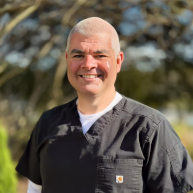Smiling man with a shaved head wearing black scrubs outdoors in daylight.
