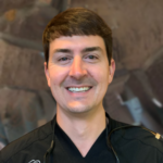 Smiling male healthcare professional wearing black scrubs with a stethoscope around his neck, in a clinical setting.