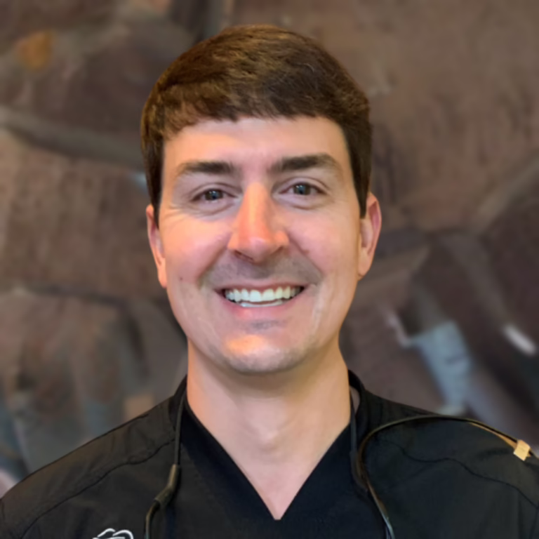Smiling male healthcare professional wearing black scrubs with a stethoscope around his neck, in a clinical setting.