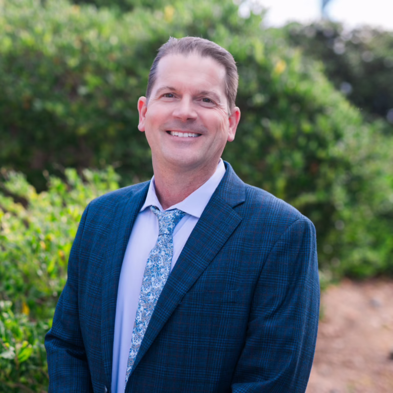 Portrait of a smiling man in a blue blazer outdoors with greenery in the background, wearing a light blue shirt and patterned tie.