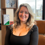 Smiling woman with shoulder-length wavy blonde hair in a black top, seated indoors near wooden shelves.