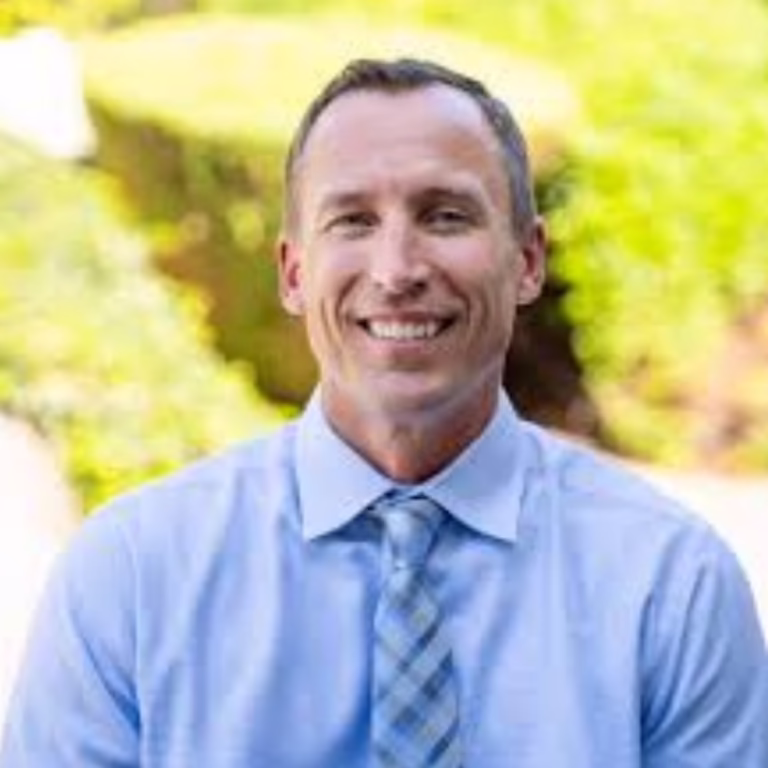Smiling man in a light blue shirt and plaid tie outdoors with greenery behind him.