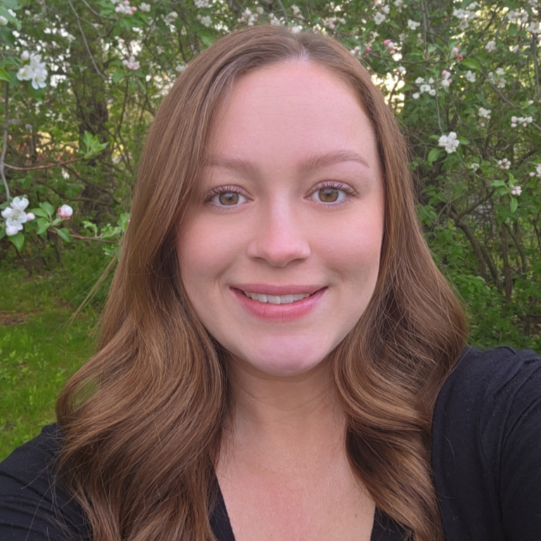 Smiling woman with long wavy brown hair outdoors among blooming white flowers in greenery.