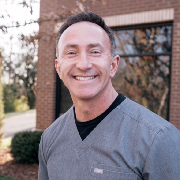 Smiling man in gray medical scrubs posing outdoors in front of a brick building.