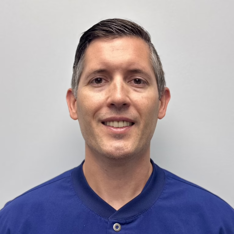 Smiling man with short dark hair wearing a blue shirt against a light gray background (head-and-shoulders portrait).
