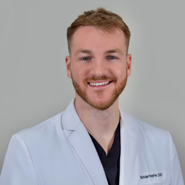 Portrait of a smiling male doctor in a white coat against a neutral gray background, looking at the camera.
