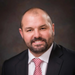 Head-and-shoulders portrait of a smiling man in a black suit, white shirt, and red striped tie against a dark studio backdrop, beard neatly trimmed.