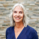 Smiling older woman with silver hair in a navy blouse standing in front of a stone wall.