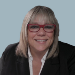 Smiling woman with blonde hair and red glasses, wearing a black blazer and white shirt, portrait against a blue background.