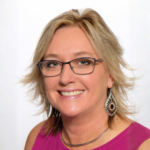 Smiling woman with short blonde hair and black glasses wearing a magenta top and large dangling earrings.