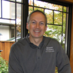 Smiling man with short gray hair in a dark embroidered shirt standing indoors by large windows.