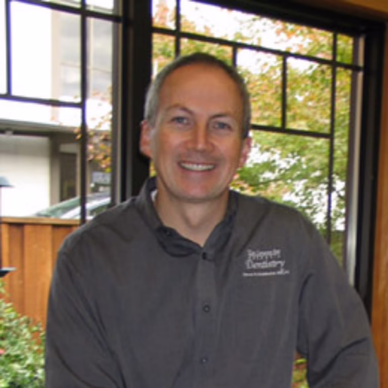 Smiling man with short gray hair in a dark embroidered shirt standing indoors by large windows.