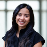 Smiling young woman with long dark hair in a black blouse, against a soft, blurred background.