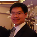 Asian man in a dark suit and patterned tie, smiling at a formal event with floral decor in the background.