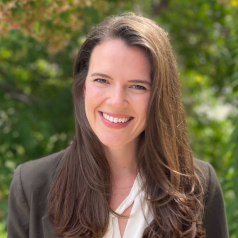 Smiling woman with long brown hair in a blazer outdoors, with green foliage behind her.