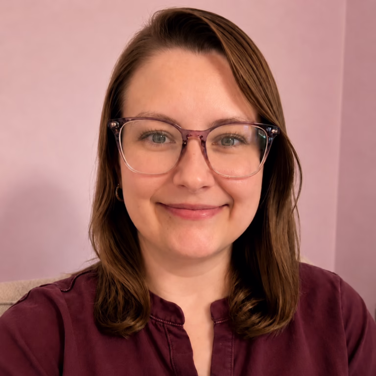Smiling woman with shoulder-length brown hair and pink-framed glasses wearing a burgundy top, in front of a pink background (self-portrait)