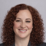 Portrait of a smiling woman with shoulder-length curly red hair, wearing a dark blazer against a neutral gray background.