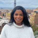 Portrait of a smiling woman with long dark hair wearing a white knit sweater, against a cityscape and sea in the distance.