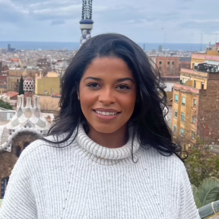 Portrait of a smiling woman with long dark hair wearing a white knit sweater, against a cityscape and sea in the distance.