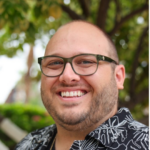 Smiling man with short beard wearing dark glasses and a black-and-white tropical print shirt outdoors.