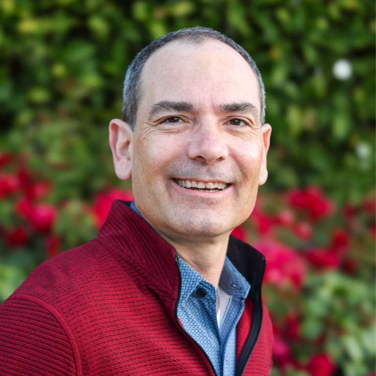 Smiling man in a red jacket outdoors with green foliage in the background (head-and-shoulders portrait)