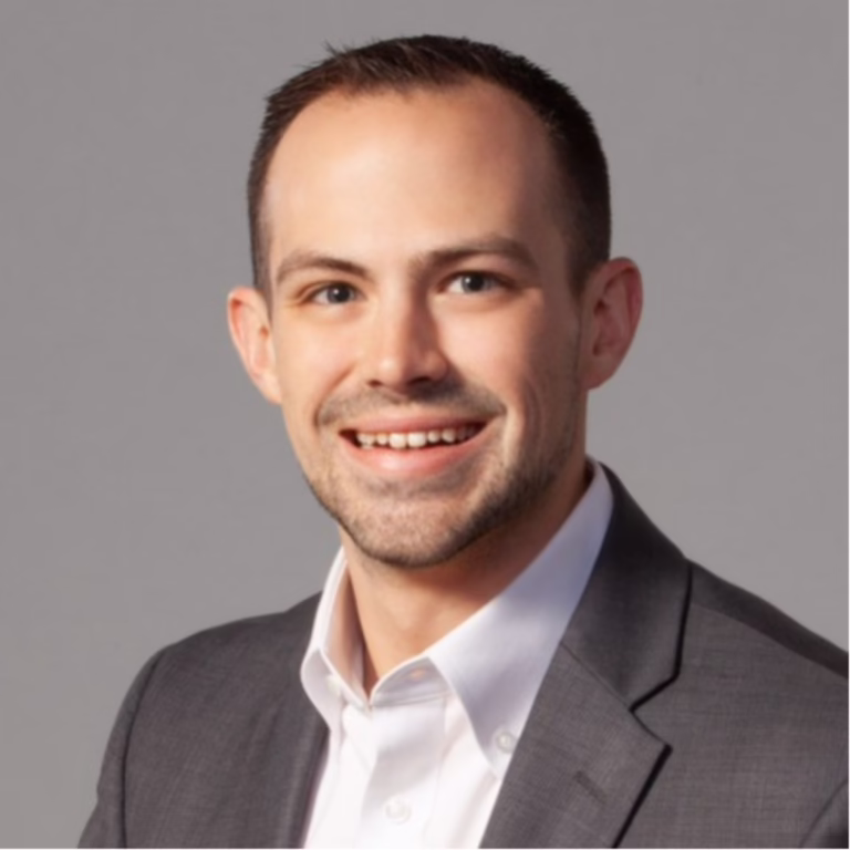 Smiling man in a gray suit and white shirt, professional headshot against a neutral background.
