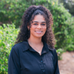 Smiling woman with curly hair wearing a dark button-up shirt, posing outdoors with green foliage in the background.