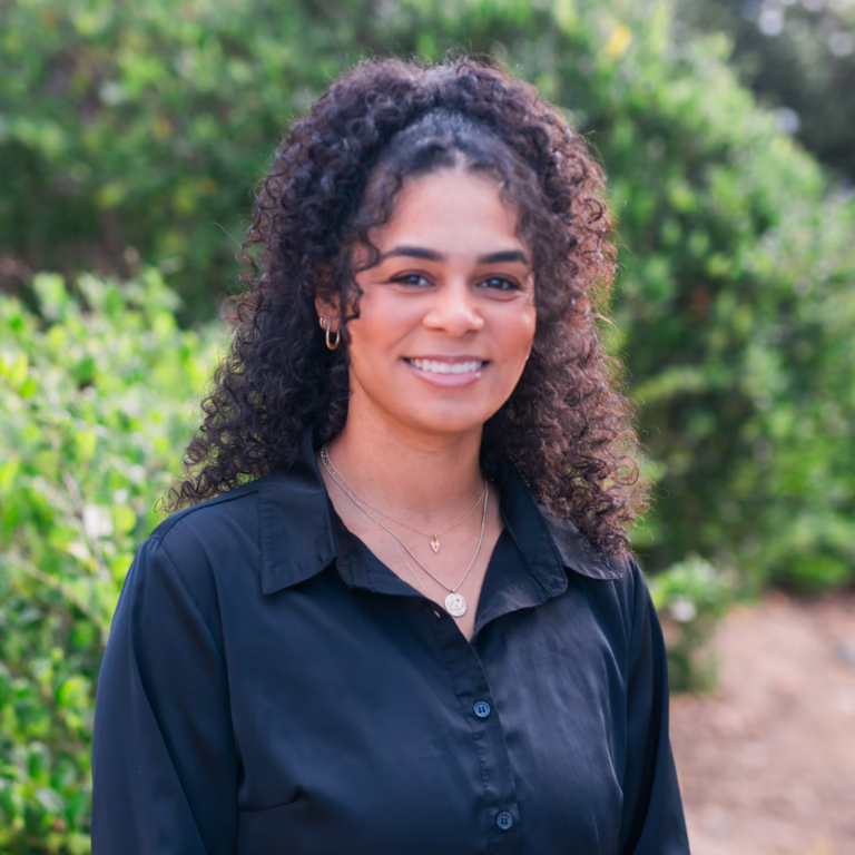 Smiling woman with curly hair wearing a dark button-up shirt, posing outdoors with green foliage in the background.