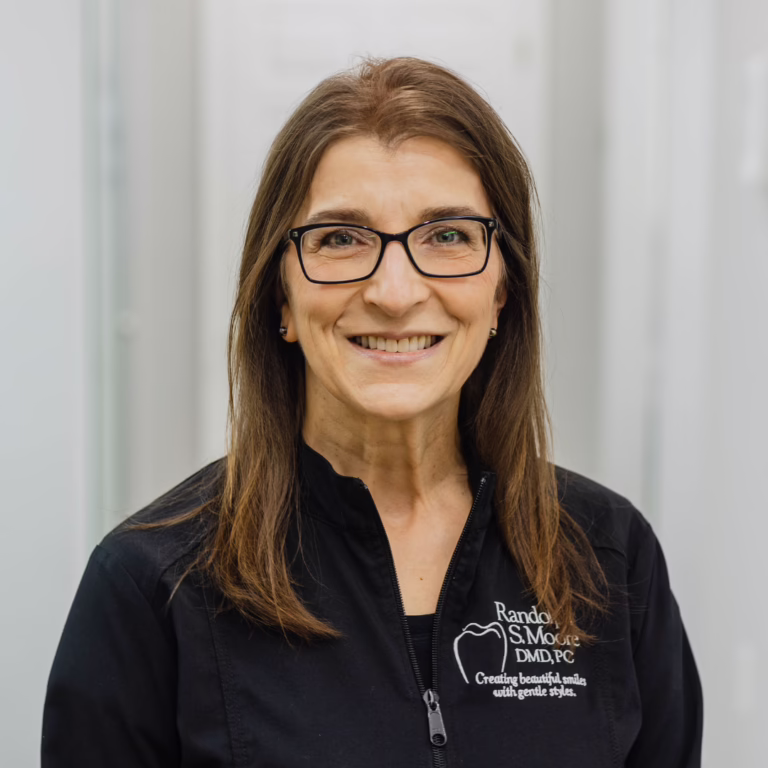 Smiling female dentist wearing glasses and a black zip-up jacket with a dental logo.