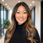 Portrait of a smiling woman with long, wavy balayaged hair wearing a black turtleneck, in a bright office hallway.