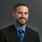 Professional headshot: smiling man in a black suit, blue shirt, and plaid tie against a gray backdrop.