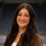 Portrait of a smiling woman with long dark hair, wearing a brown corduroy jacket and a pendant necklace outdoors.