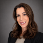 Professional headshot of a smiling woman with brown hair, wearing a black blazer against a gray studio backdrop.