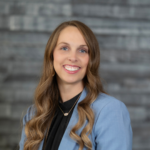Smiling woman with long wavy hair in a blue blazer and black blouse against a grey brick wall.