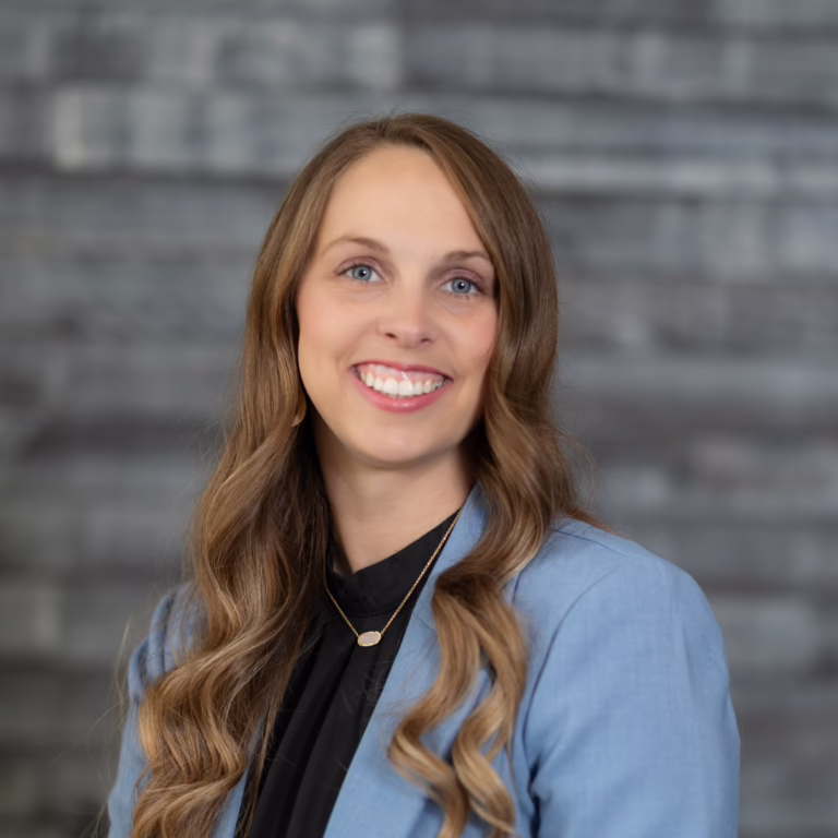 Smiling woman with long wavy hair in a blue blazer and black blouse against a grey brick wall.
