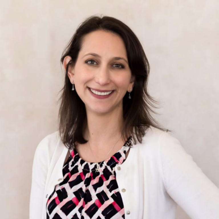 Portrait of a smiling woman with shoulder-length dark hair wearing a white cardigan over a pink-black patterned blouse, neutral background.