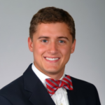 Professional headshot of a smiling young man in a navy suit and red bow tie against a gray backdrop.
