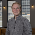 Portrait of a smiling man in a gray quarter-zip sweater indoors with large windows behind him, suggesting a corporate headshot.