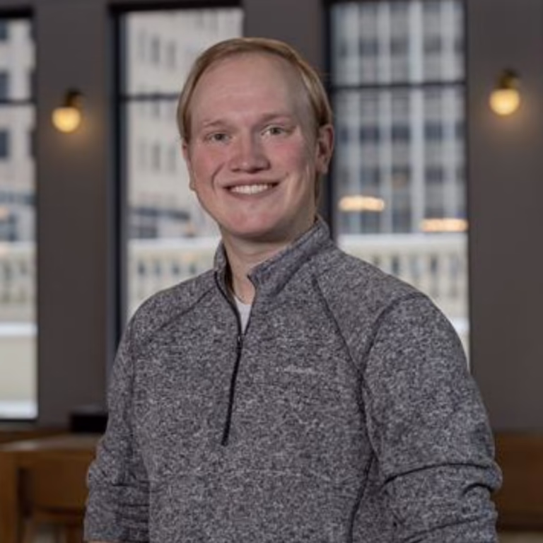 Portrait of a smiling man in a gray quarter-zip sweater indoors with large windows behind him, suggesting a corporate headshot.