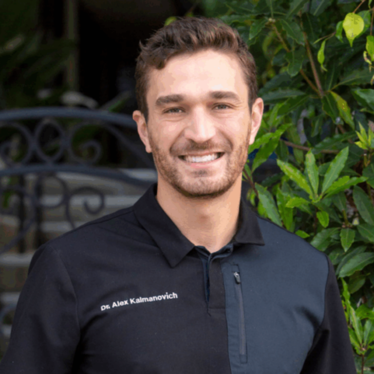 Portrait of a smiling man in a black polo shirt, head and shoulders, outdoors with green foliage in the background.