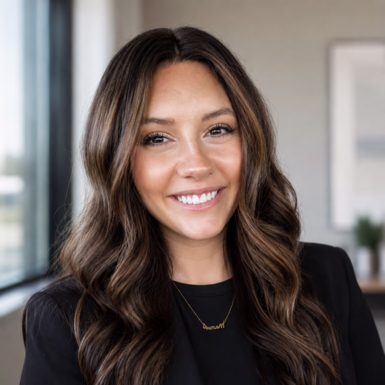 Smiling woman with long wavy brown hair in a black blazer, indoors in a sunny office setting.