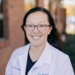 Portrait of a smiling female doctor wearing glasses and a white coat outdoors.