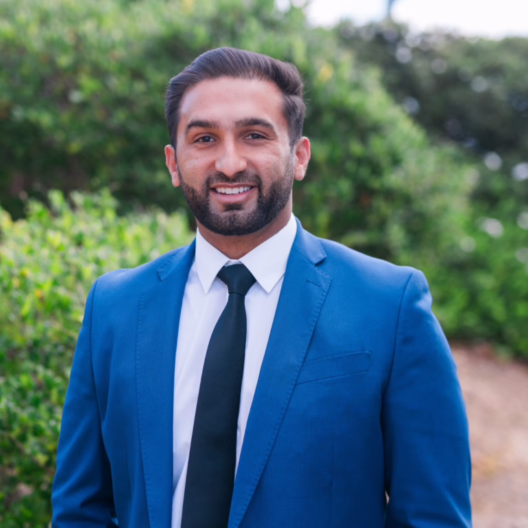 Smiling man in a blue suit and black tie, standing outdoors with greenery in the background (portrait).