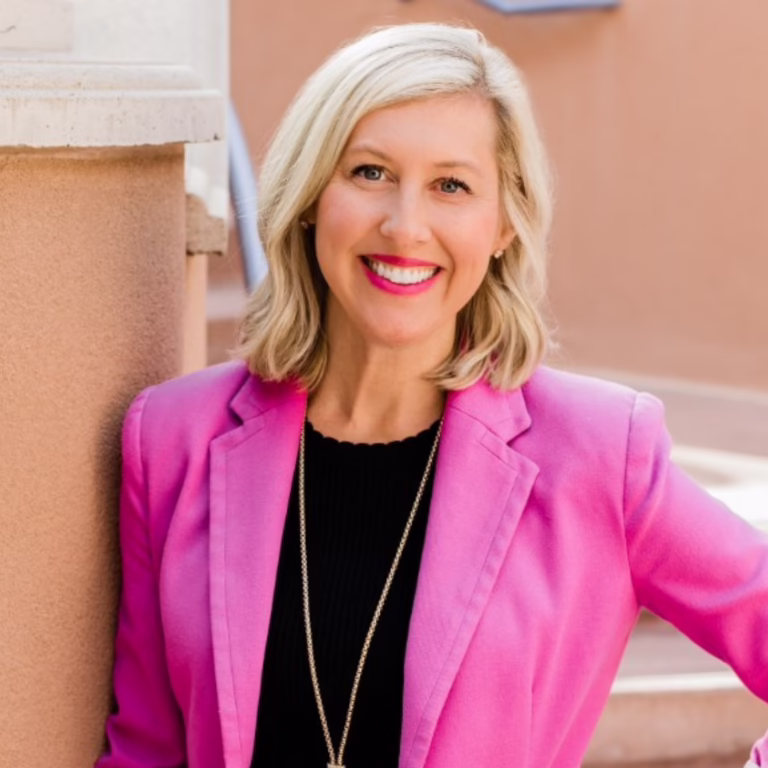 Smiling woman with short blonde hair in a pink blazer and black top, standing outdoors beside a wall.