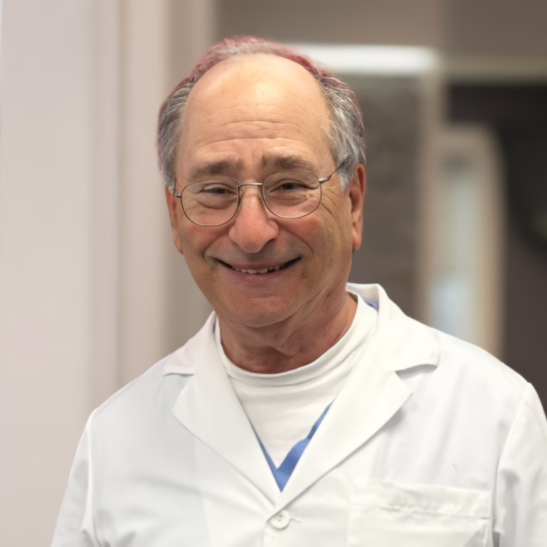 Smiling older man in a white lab coat and glasses in a medical setting.