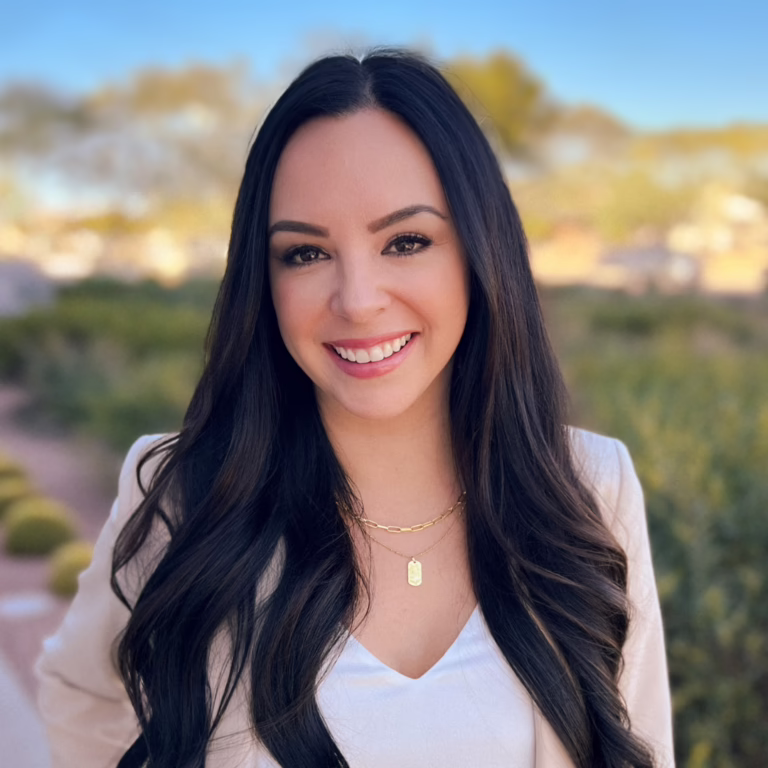 Portrait of a smiling woman with long dark hair outdoors, wearing a white top and beige blazer with gold layered necklaces.
