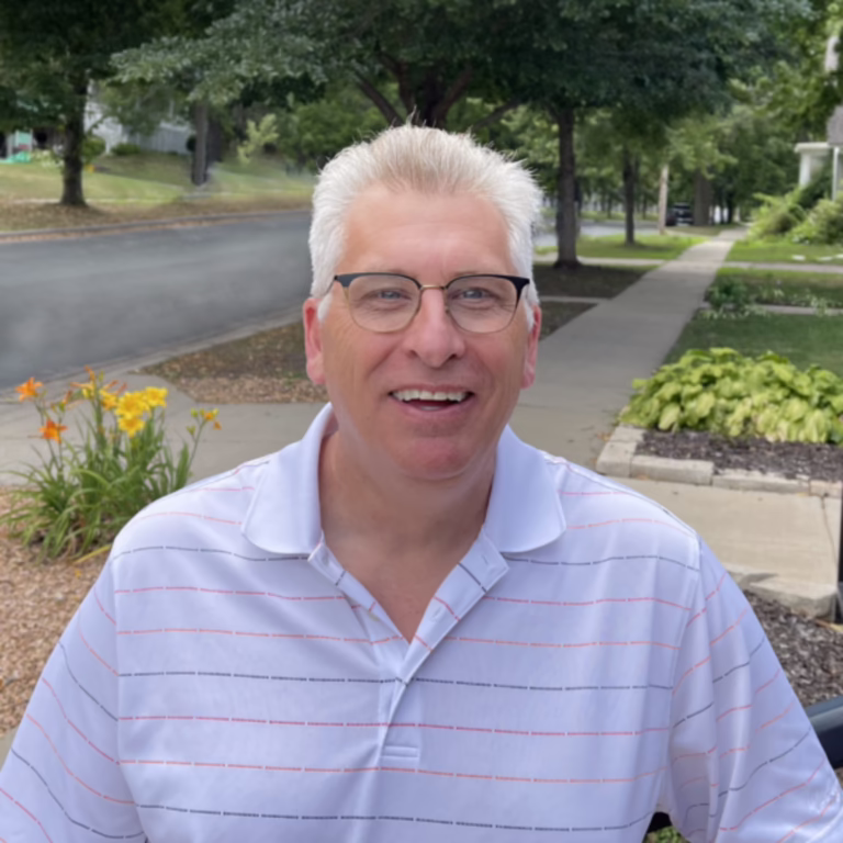 Smiling man with gray hair and glasses standing on a sidewalk outdoors, wearing a white striped polo shirt with red and blue lines, greenery and flowers in the background.