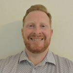 Portrait of a smiling man with red hair and a red beard, wearing a patterned button-down shirt against a light background.